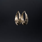 Presenting the PROMETHEUS Medium Biomechanical Hoop Earrings in Gold by Kaya Movement: these unisex earrings feature an intricate, gold-colored design with detailed, curved and woven elements that evoke biomechanical artistry. The 18 gauge hoops are set against a plain black background and have a clasp at the top for secure fastening.