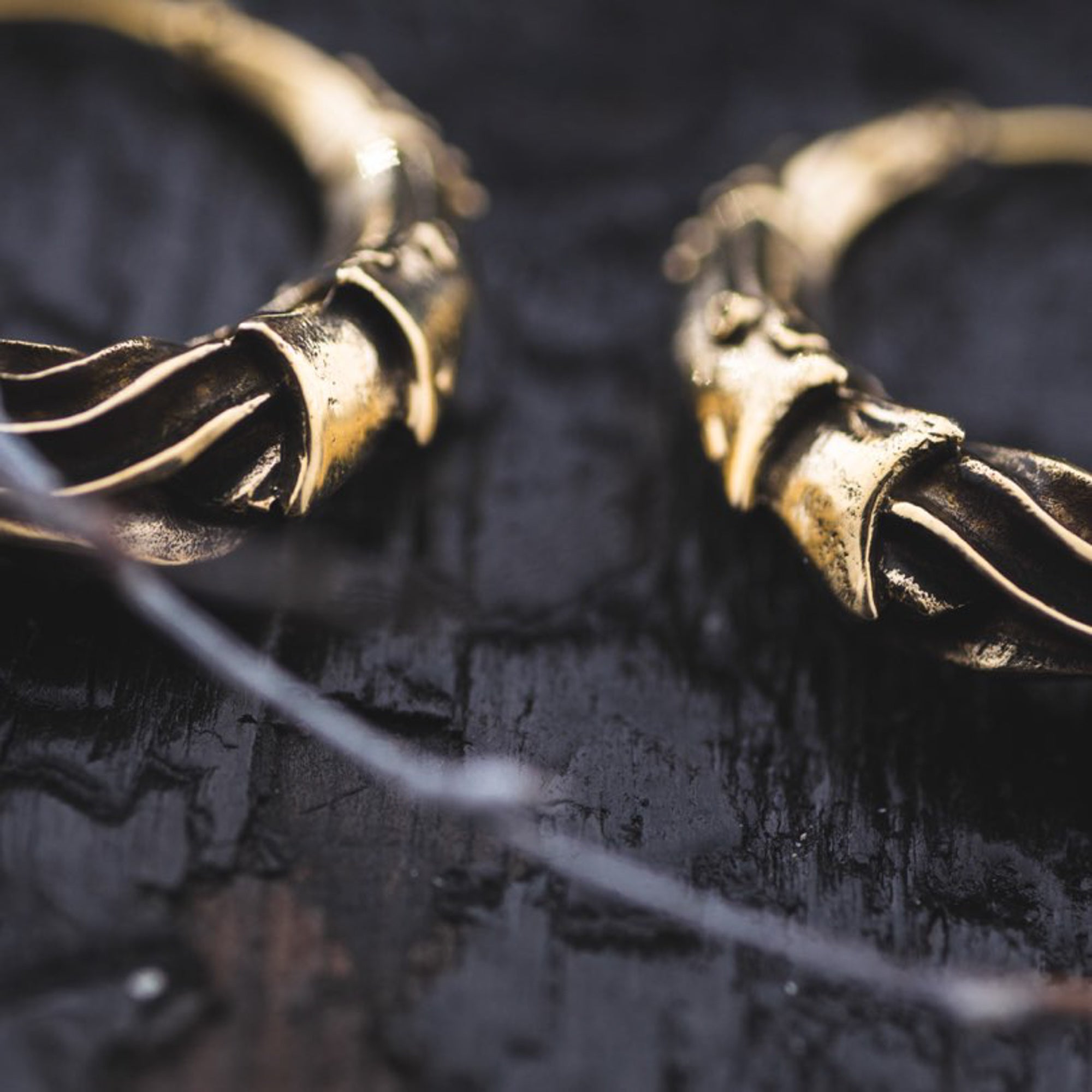 Close-up view of two intricately designed PROMETHEUS Small Cyborg Hoop Earrings in Gold placed adjacent to each other against a rough, dark wooden surface. The earrings feature detailed, textured patterns that give them a unique and luxurious appearance, reminiscent of post-apocalyptic body jewelry by Kaya Movement.