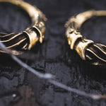 Close-up view of two intricately designed PROMETHEUS Small Cyborg Hoop Earrings in Gold placed adjacent to each other against a rough, dark wooden surface. The earrings feature detailed, textured patterns that give them a unique and luxurious appearance, reminiscent of post-apocalyptic body jewelry by Kaya Movement.