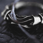 A close-up shot of a textured, silver earring from Kaya Movement's PROMETHEUS Small Cyborg Hoop Earrings collection is placed on a dark background. The earring has an intricate, wavy design and is partially covered by a thin, curved twig. The lighting emphasizes the details and contours of the 925 silver piece, making it reminiscent of post-apocalyptic body jewelry.