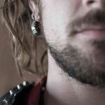 Close-up photograph of a person's face focusing on the lower half. The individual has a beard, a mustache, and is wearing PROMETHEUS Small Cyborg Hoop Earrings 925 Silver | 18 gauge from Kaya Movement. Their hair is wavy and hangs over their ear. The background is blurred, drawing attention to the facial details and earrings.