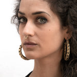 A woman with curly dark hair and piercing blue eyes stares directly at the camera. She wears a nose ring and large, ornate QUETZALCOATL Oversized Snake Spine Hoop Earrings in Gold by Kaya Movement, which have a textured pattern that gives off a sci-fi statement earrings vibe. The background is a plain, light color, allowing her features to stand out prominently.