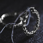 A close-up shot captures a pair of Quetzalcoatl Oversized Snake Spine Hoop Earrings in Silver White Brass by Kaya Movement, resting on a dark textured surface. One earring is in sharp focus, highlighting its intricate design and detailed pattern, while the other remains blurry in the background. Thin light strands drape across the scene, adding to the visual intrigue.