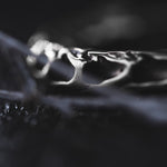 Close-up of a textured, silver-colored earring from Kaya Movement's Quetzalcoatl Oversized Snake Spine Hoop Earrings in Silver White Brass. The intricate design brings to mind post-apocalyptic body jewelry. The dark and blurry background creates a dramatic contrast, highlighting the earring's details and its fluid, organic pattern.