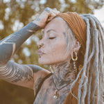A person with long, grey dreadlocks and extensive tattoos on their face, neck, and arm is pictured outdoors. They wear a brown headwrap and post-apocalyptic body jewelry including multiple piercings and QUETZALCOATL Oversized Snake Spine Hoop Earrings in Gold (14 gauge) by Kaya Movement. Their eyes are closed, and one hand rests on their forehead, set against a backdrop of blurred foliage.
