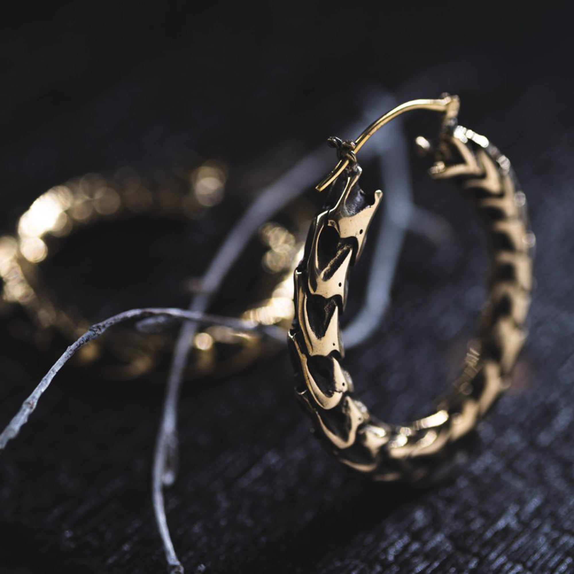 Close-up image of intricate QUETZALCOATL Oversized Snake Spine Hoop Earrings in Gold by Kaya Movement. They feature a textured, braided design and are placed on a dark, textured surface with frayed white string partially visible in the background. The soft lighting highlights the fine details of these sci-fi statement earrings.
