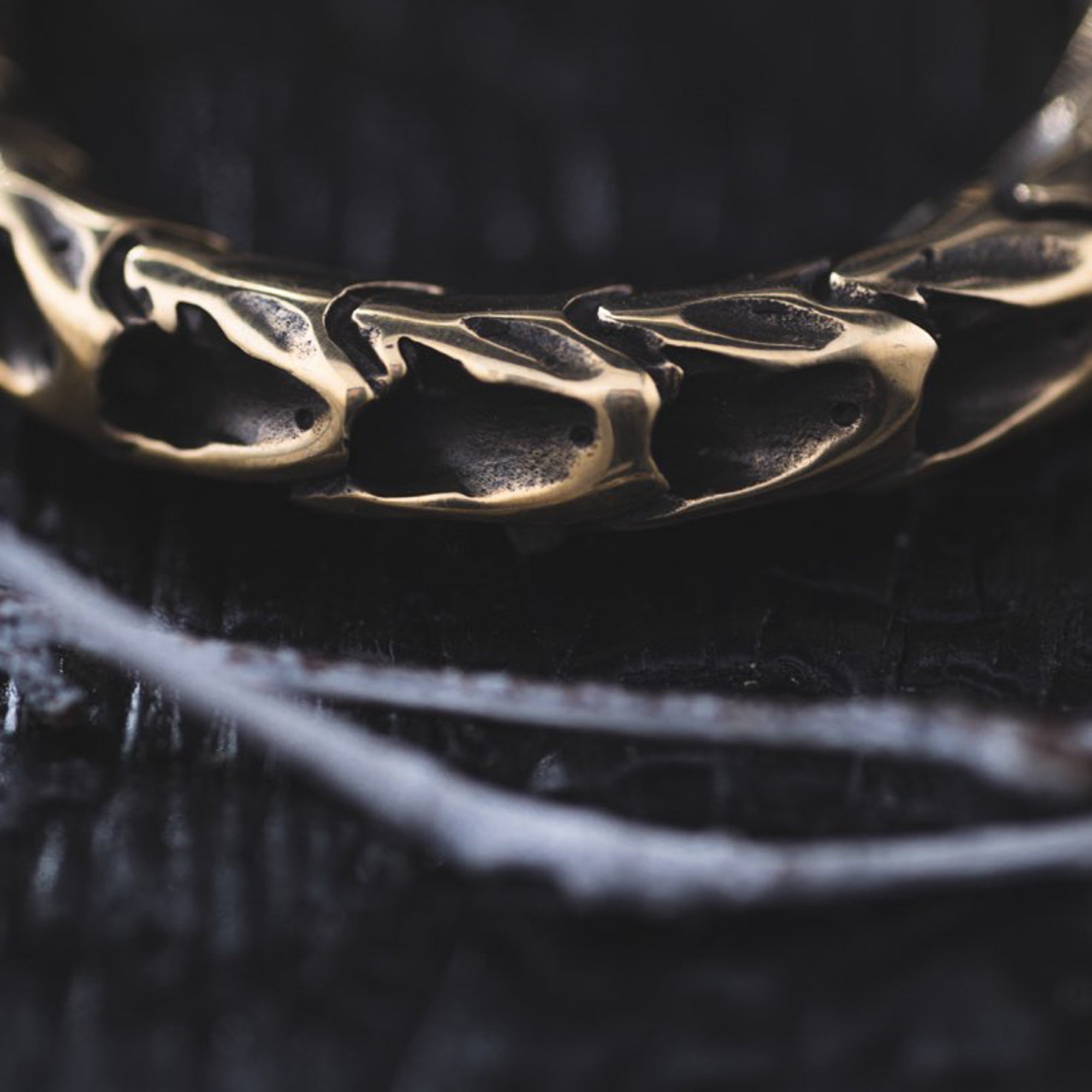 A close-up image reveals an intricately designed QUETZALCOATL Oversized Snake Spine Hoop Earring in Gold by Kaya Movement, showcasing organic patterns that evoke intertwined skeletal forms. The dark and textured background enhances the mysterious and moody atmosphere, reminiscent of post-apocalyptic body jewelry. Thin, white branches are faintly visible at the bottom of the frame.