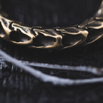 A close-up image reveals an intricately designed QUETZALCOATL Oversized Snake Spine Hoop Earring in Gold by Kaya Movement, showcasing organic patterns that evoke intertwined skeletal forms. The dark and textured background enhances the mysterious and moody atmosphere, reminiscent of post-apocalyptic body jewelry. Thin, white branches are faintly visible at the bottom of the frame.