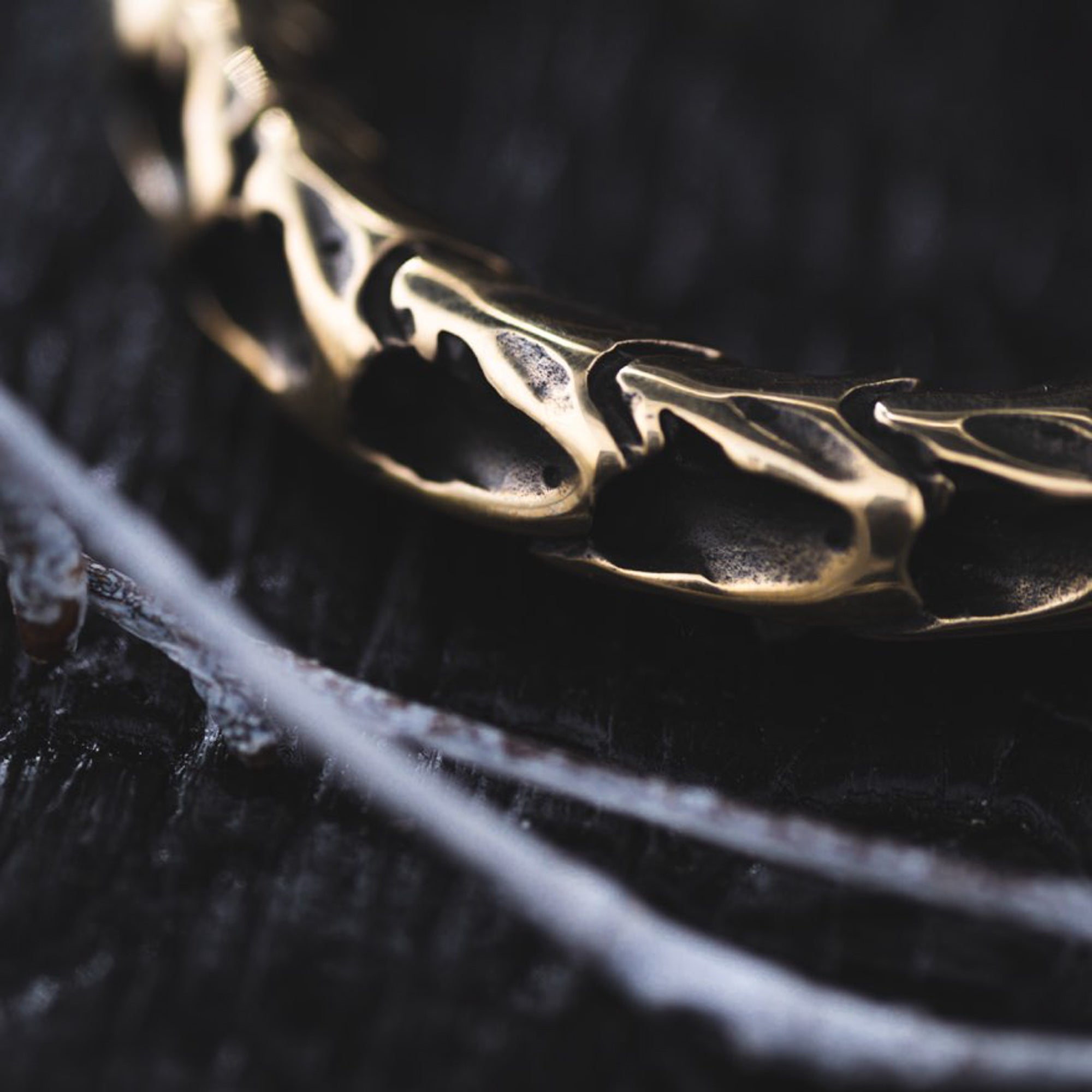 Close-up image of the QUETZALCOATL Oversized Snake Spine Hoop Earrings in Gold by Kaya Movement, showcasing their unique, intricate design on a textured dark surface. The earrings feature a rugged, organic pattern reminiscent of post-apocalyptic body jewelry, set against what appears to be charred or heavily textured wood.