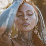 A person with freckles and tattoos poses with one arm raised, eyes closed, and adorned in post-apocalyptic body jewelry. They are wearing Kaya Movement's QUETZALCOATL Oversized Snake Spine Hoop Earrings in Gold (14 gauge), along with several facial piercings such as a septum ring, nose studs, and a lip ring. The background is softly lit and slightly blurred.