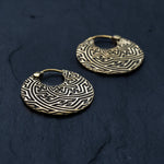 A pair of SAYA Small Tribal Sayagata Disc Hoop Earrings in Gold by Kaya Movement, featuring intricate maze-like Sayagata patterns, rests on a dark, textured surface. These 18 gauge gold earrings boast a rich, antique appearance and a small hinge closure at the top, making them the perfect boho statement jewelry.