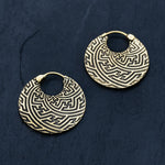 The SAYA Small Tribal Sayagata Disc Hoop Earrings in Gold by Kaya Movement are crafted from gold brass and feature an intricate Sayagata pattern on a dark, textured surface. These boho statement pieces have a maze-like design and are secured with a small clasp mechanism at the top for easy fastening to the ear.