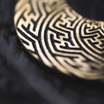 Close-up image of SAYAMOON Tribal Sayagata Seamless Hoop Disc Earrings in Gold by Kaya Movement, showcasing intricate, maze-like patterns etched into their metallic surface. Resembling tribal round disc earrings, these 12 gauge accessories have a gold sheen, and their texture stands out against a dark, blurred background.