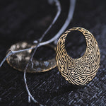 A pair of Kaya Movement's SAYAMOON Tribal Sayagata Seamless Hoop Disc Earrings in Gold (12 gauge) are displayed on a dark surface. One earring is upright, while the other lies flat. Thin twigs are also part of the arrangement, adding a natural contrast to the ornate jewelry.