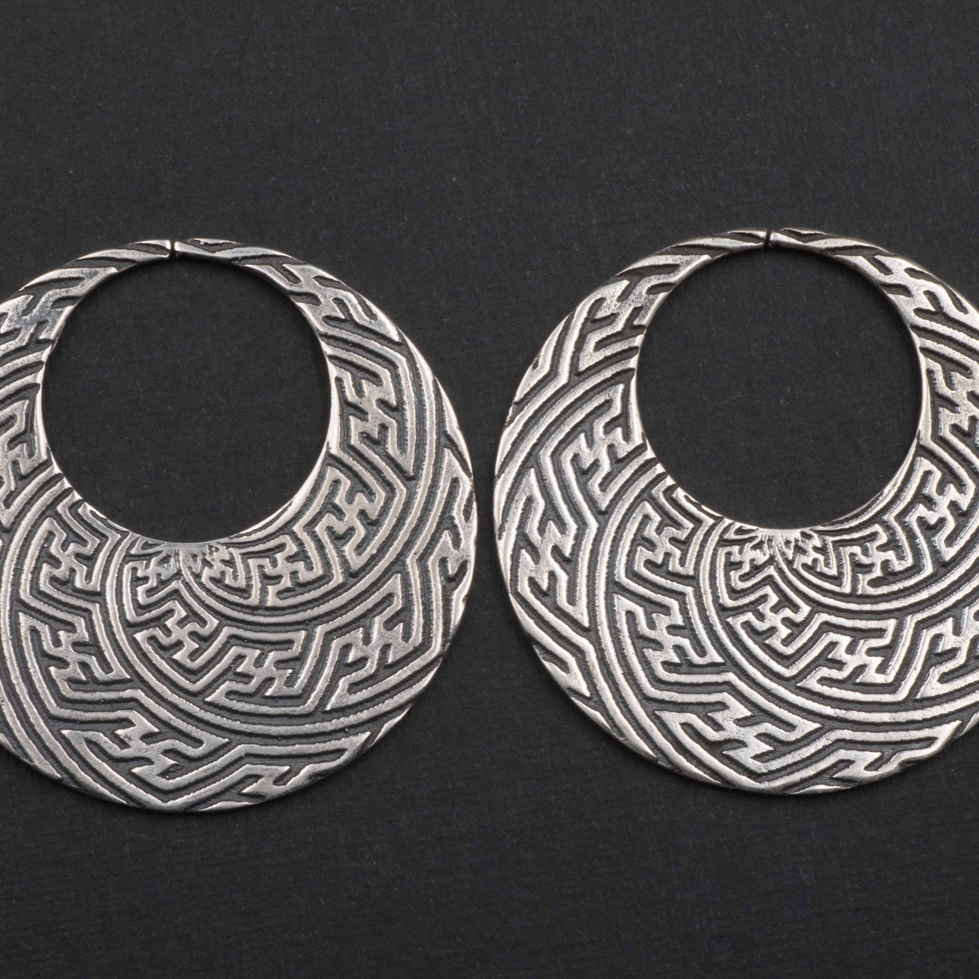 The SAYAMOON Tribal Sayagata Hoop Disc Earrings in 925 Silver by Kaya Movement feature large, circular Sterling silver earrings with intricate, geometric patterns etched into the metal on a solid black background. These detailed and textured designs are reminiscent of tribal body jewelry. They are constructed in a 12 gauge size.