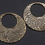 The SAYAMOON Tribal Sayagata Seamless Hoop Disc Earrings in Gold | 12 gauge by Kaya Movement lay on a dark surface, showcasing an intricate, repeating geometric pattern etched into their metallic surface. These boho statement earrings incorporate interlocking shapes reminiscent of a maze or labyrinth.
