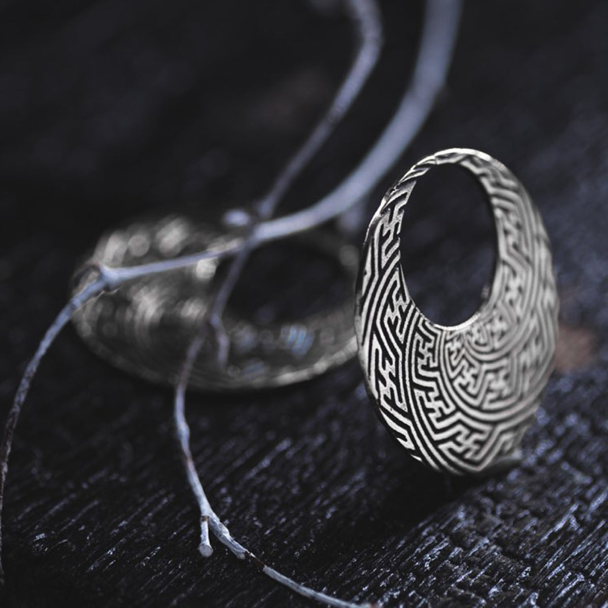 A close-up of the SAYAMOON Tribal Sayagata Hoop Disc Earrings in 925 Silver by Kaya Movement, featuring an intricately designed maze-like pattern, sits on a dark surface. Nearby, a thin, twisted branch lies partially blurred in the background, adding a natural element to this elegant piece of tribal body jewelry.