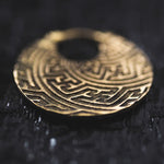 Close-up of a Kaya Movement SAYA Small Tribal Sayagata Disc Hoop Earring in Gold with intricate, maze-like engravings reminiscent of boho statement jewelry. The earring lies on a dark, textured surface, highlighting its detailed design and Sayagata pattern. The lighting creates a soft reflection on the earring’s surface, emphasizing its craftsmanship.