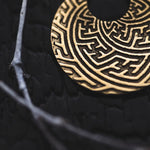 Close-up of a textured gold earring featuring intricate Sayagata patterns from the SAYA Small Tribal Sayagata Disc Hoop Earrings collection by Kaya Movement, placed on a dark surface. A gray branch partially overlaps the earring, adding contrast to this boho statement jewelry piece.