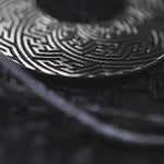 Close-up of tribal-inspired SAYAMOON Tribal Sayagata Hoop Disc Earrings in 925 Silver by Kaya Movement, featuring intricate maze-like engravings. The pendant earrings are displayed with a dark string slightly out of focus, against a dark background that highlights the detailed texture.