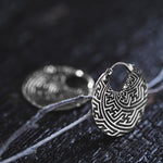 A close-up of the Kaya Movement's SAYA Small Tribal Sayagata Endless Hoop Earrings in 925 sterling silver, showcasing intricate maze-like patterns. The boho statement earrings are placed on a dark, textured surface, with one earring sharply focused in the foreground and the other artfully blurred in the background.