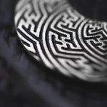 Close-up of the SAYAMOON Tribal Sayagata Hoop Disc Earrings in 925 Silver by Kaya Movement, showcasing their intricate, maze-like patterns on a metallic circular surface. The design is set against a blurred, dark background, highlighting the texture and detail of the engraving.