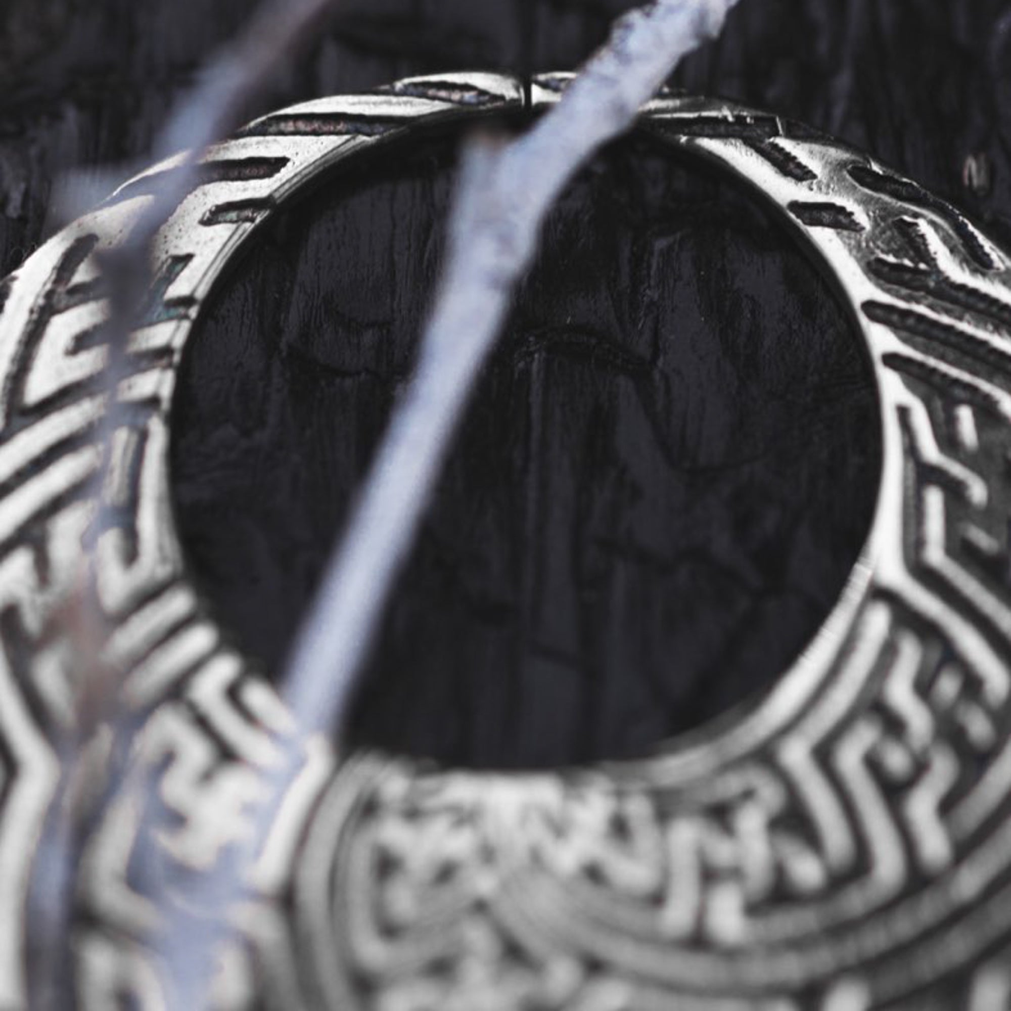 A close-up image of a pair of intricately designed SAYAMOON Tribal Sayagata Hoop Disc Earrings in 925 Silver by Kaya Movement, featuring geometric patterns. The earrings, reminiscent of tribal body jewelry, hang among blurred, translucent branches or wires with a dark, textured background that highlights the fine details of the metalwork and their 12 gauge thickness.