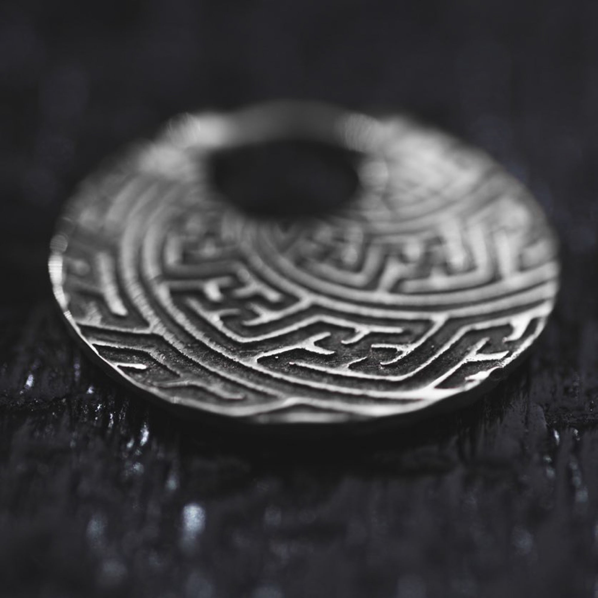 Close-up of a SAYA Tribal Sayagata Endless Hoop Earring in silver, lying flat on a dark, textured surface. The 18 gauge earring from Kaya Movement, reminiscent of dark tribal body jewelry, features intricate embossed patterns with interlocking geometric shapes, creating a visually striking labyrinth-like design. There's a small hole at the center of the earring.