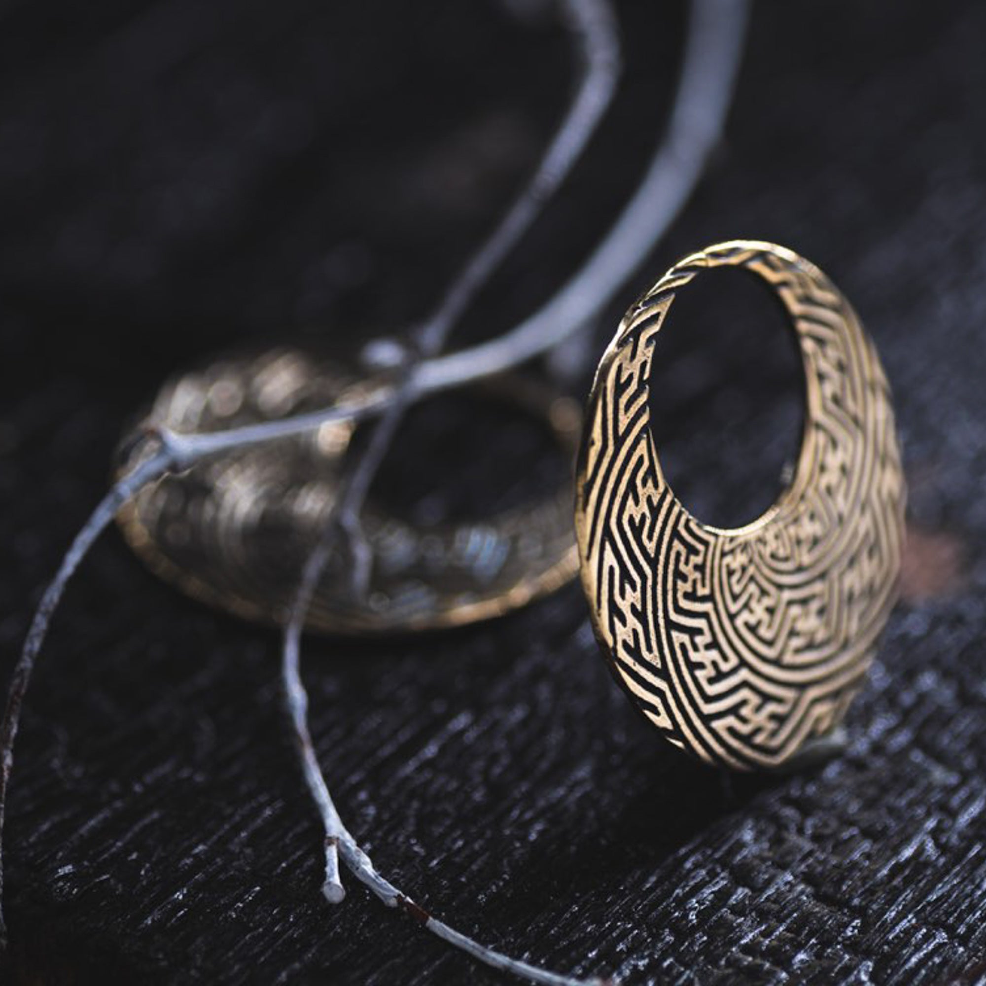 A close-up of the SAYAMOON Tribal Sayagata Seamless Hoop Disc Earrings in Gold by Kaya Movement, showcasing its circular shape and intricate, maze-like patterns. The 12-gauge gold brass earring rests on a dark surface, with delicate, thin branches intertwined nearby.