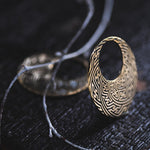 A close-up of the SAYAMOON Tribal Sayagata Seamless Hoop Disc Earrings in Gold by Kaya Movement, showcasing its circular shape and intricate, maze-like patterns. The 12-gauge gold brass earring rests on a dark surface, with delicate, thin branches intertwined nearby.