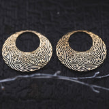 SAYAMOON Tribal Sayagata Seamless Hoop Disc Earrings in Gold from Kaya Movement with intricate, maze-like patterns are set against a dark, textured background. A slender twig lies diagonally at the bottom of the image.