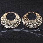 SAYAMOON Tribal Sayagata Seamless Hoop Disc Earrings in Gold from Kaya Movement with intricate, maze-like patterns are set against a dark, textured background. A slender twig lies diagonally at the bottom of the image.