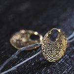 A pair of SAYA Small Tribal Sayagata Disc Hoop Earrings in Gold by Kaya Movement rest on a dark, textured surface. The 18 gauge earrings feature an intricate labyrinthine Sayagata pattern, giving them an ornate and vintage appearance. One earring stands upright while the other lies flat in the background.