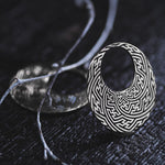 A close-up shot of an intricately designed SAYAMOON Tribal Sayagata Hoop Disc Earring in 925 Silver from Kaya Movement, placed on a dark textured surface. The earring showcases an elaborate labyrinth pattern reminiscent of tribal body jewelry, and a thin, curved branch is de-focused in the background, adding contrast to the composition.