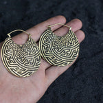 A hand holds the SAYAGATA Oversized Tribal Hoop Disc Earrings in Gold by Kaya Movement. These large earrings, made of brass, feature intricate maze-like patterns. The dark and textured background contrasts with the detailed design of these boho statement pieces.