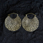 Displayed against a dark, textured background are a pair of SAYAGATA Oversized Tribal Hoop Disc Earrings in Gold from Kaya Movement. These 16-gauge earrings have an antique finish and hook closure, showcasing intricate labyrinthine patterns and a detailed, geometric boho design.