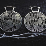 Introducing the SEIGAIHA Oversized Japanese Geometry Disc Earrings by Kaya Movement. These 16 gauge, intricate silver earrings feature a wave pattern design set against a dark background with thin silver wires attached at the top for wearing. A light branch is also visible in the foreground, enhancing their boho statement appeal.