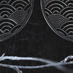 Close-up of a black surface with a pattern of overlapping arches resembling waves. Intricate designs are visible on two circular metallic objects, which are the SEIGAIHA Oversized Japanese Geometry Disc Earrings in Silver by Kaya Movement. The background appears dark, with a frosty white twig at the bottom of the image.