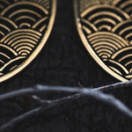 Close-up of the SEIGAIHA Oversized Japanese Geometry Disc Earrings in Gold by Kaya Movement, showcasing their intricate curved line patterns that resemble waves. The black and gold unisex statement earrings are placed on a dark surface, with a blurred element resembling a thread or wire in the foreground. The design is highly detailed and elegant.