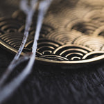 Close-up of the SEIGAIHA Oversized Japanese Geometry Disc Earrings in Gold by Kaya Movement, featuring an intricately patterned wave-like design reminiscent of tribal jewelry, partially obscured by two out-of-focus white branches in the foreground. The earrings rest on a dark, textured surface.