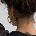The image shows the back of a person’s head and neck, featuring SEIGAIHA Oversized Japanese Geometry Disc Earrings in Gold by Kaya Movement. The circular earrings have an intricate wave pattern and are 16 gauge in size. The person has dark, curly hair pulled back, revealing a small tattoo on the nape of their neck. These unisex statement earrings blend tribal jewelry and boho aesthetics seamlessly.