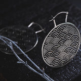 Close-up of the SEIGAIHA Oversized Japanese Geometry Disc Earrings in Silver by Kaya Movement, displayed on a dark textured surface. The intricately designed 16-gauge earrings feature a repeating pattern of overlapping semicircles and are resting near a slender tree branch. The dim lighting emphasizes the detailed craftsmanship of the earrings.