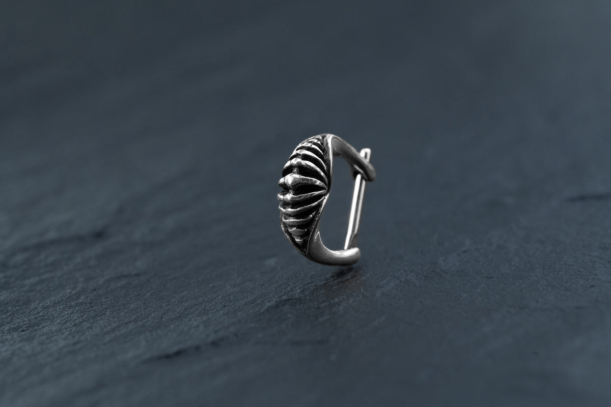 A silver "DIARA Biomechanical Clicker Hoop Septum Nose Ring in Silver | 16 gauge" from Kaya Movement rests on a dark, slate surface. The piece displays smooth curves and detailed engraving that highlight its elegant and intricate craftsmanship—reminiscent of the fine artistry seen in unisex 16 gauge nose piercing designs.