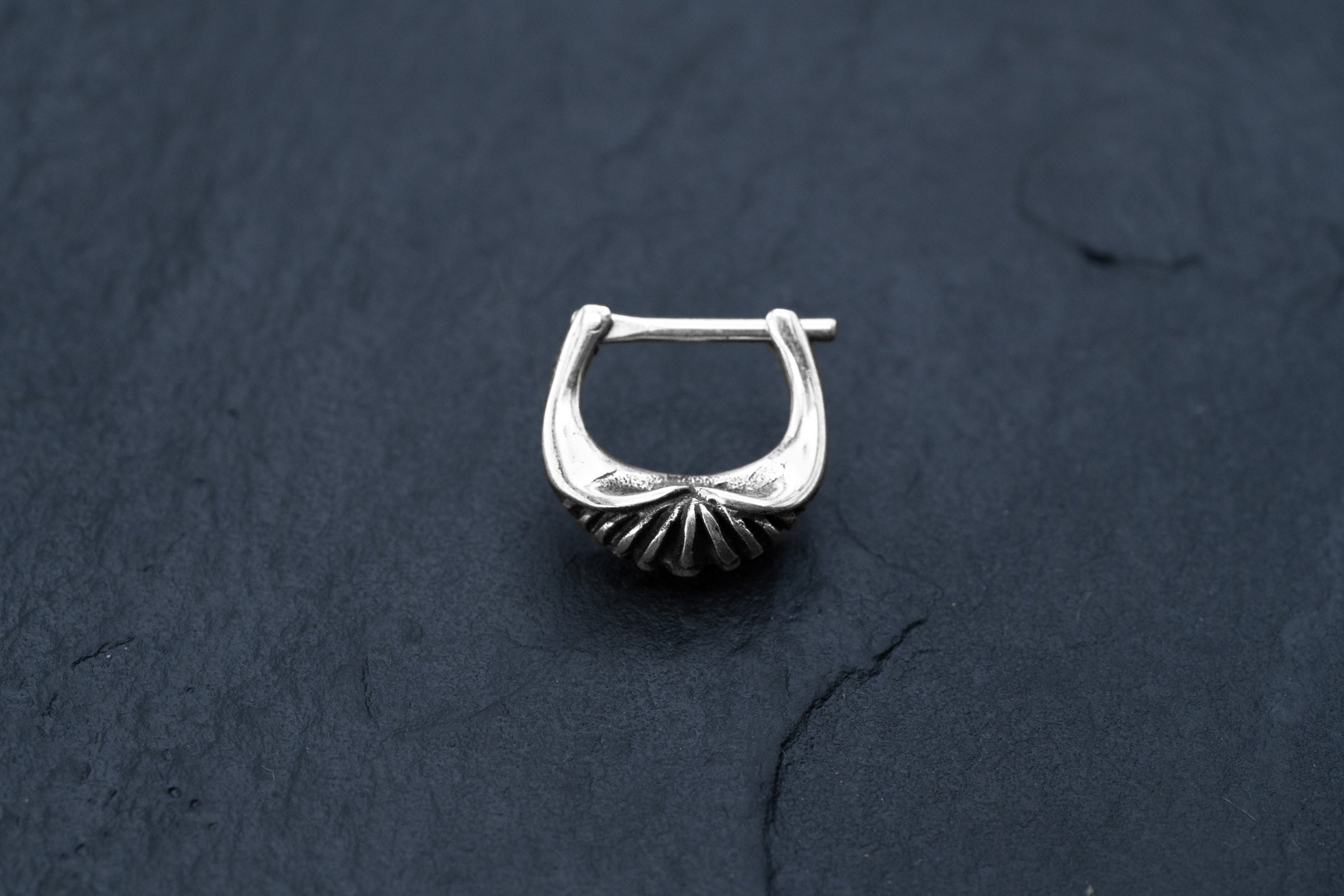 The DIARA Biomechanical Clicker Hoop Septum Nose Ring in Silver by Kaya Movement is showcased against a dark, textured background. This 16 gauge earring, with its intricate shell-like design at the base and horseshoe shape complemented by a bar top for fastening, exudes elegance and sophistication.