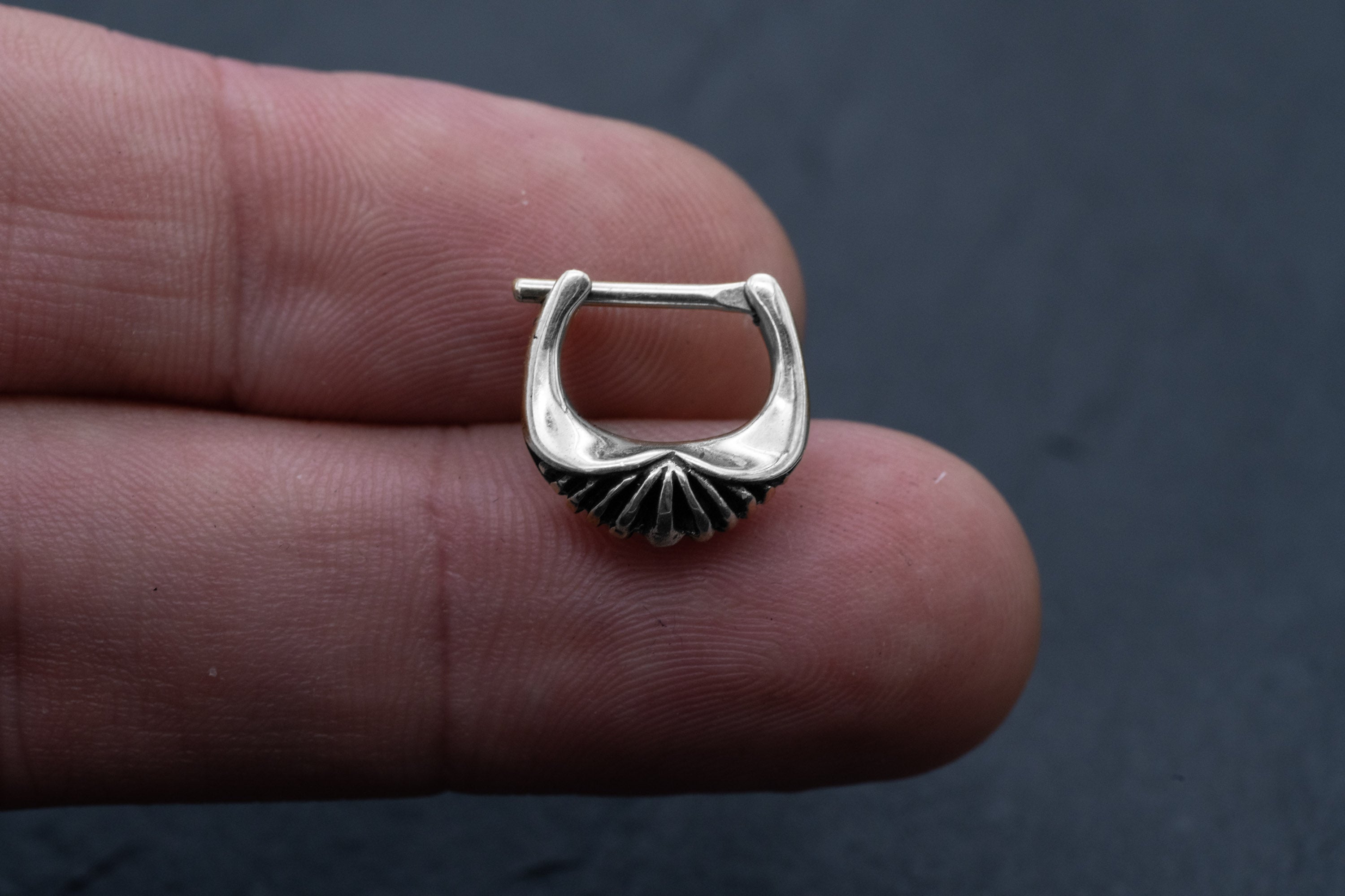 A close-up image of a hand holding the DIARA Biomechanical Clicker Hoop Septum Nose Ring in Silver by Kaya Movement. Crafted from 925 silver and designed as a 16 gauge piece, this stunning septum ring, resembling a curved fan or shell with ridges and a smooth finish, stands out against the black background, which highlights the intricate details of this exquisite body jewelry.