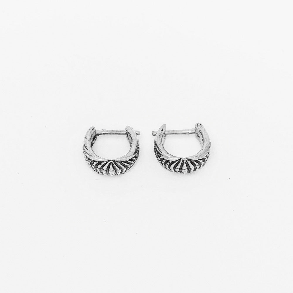 The Darma Biomechanical Clicker Hoop Septum Nose Ring in Silver by Kaya Movement is a pair of small, intricately engraved, silver hoops featuring lines extending from the base to the top. Reminiscent of post-apocalyptic body jewelry, they have a latch back closure and are displayed against a plain white background.