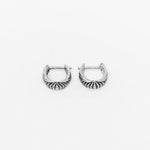 The Darma Biomechanical Clicker Hoop Septum Nose Ring in Silver by Kaya Movement is a pair of small, intricately engraved, silver hoops featuring lines extending from the base to the top. Reminiscent of post-apocalyptic body jewelry, they have a latch back closure and are displayed against a plain white background.