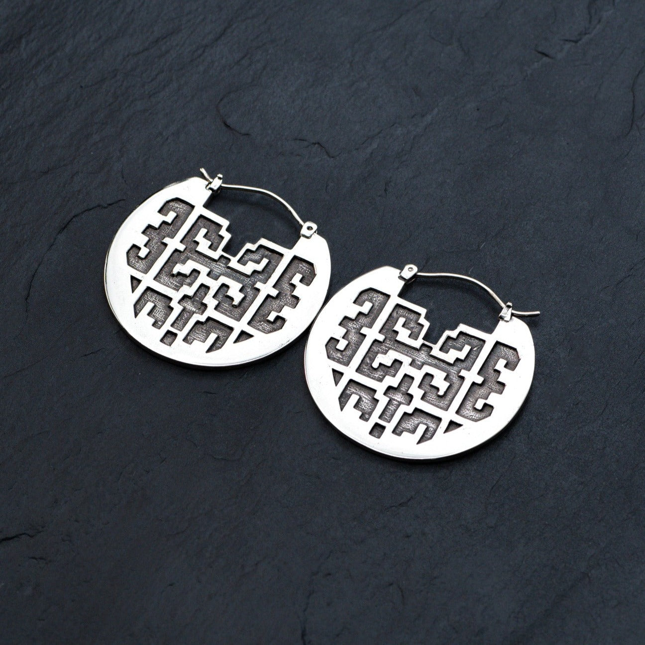 The CONIBOS Tribal Shipibo Hoop Disc Earrings in Silver from Kaya Movement, made with 16 gauge silver brass, showcase a circular design with intricate cut-out patterns. Placed on a dark textured surface, these earrings are perfect for dark boho body jewelry enthusiasts.