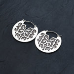 The CONIBOS Tribal Shipibo Hoop Disc Earrings in Silver from Kaya Movement, made with 16 gauge silver brass, showcase a circular design with intricate cut-out patterns. Placed on a dark textured surface, these earrings are perfect for dark boho body jewelry enthusiasts.