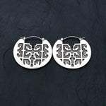 Close-up of the CONIBOS Tribal Shipibo Hoop Disc Earrings in Silver by Kaya Movement against a dark textured background. These 16 gauge earrings feature an intricate cut-out geometric pattern, creating a symmetric and ornate design that evokes the essence of dark boho body jewelry.